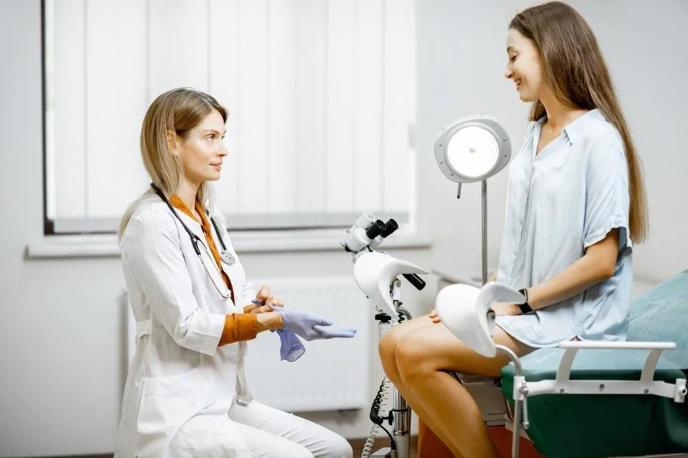 A gynecologist talking with a patient seated on an exam chair beside a colposcope