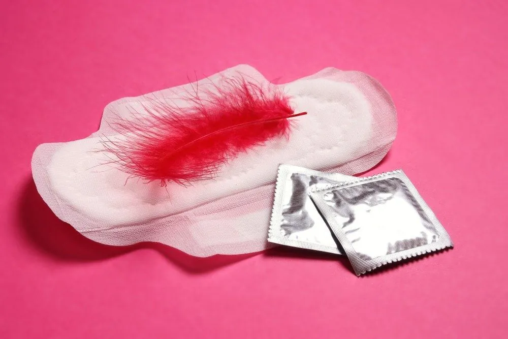 A pad with a red feather on it next to two condoms on a pink background