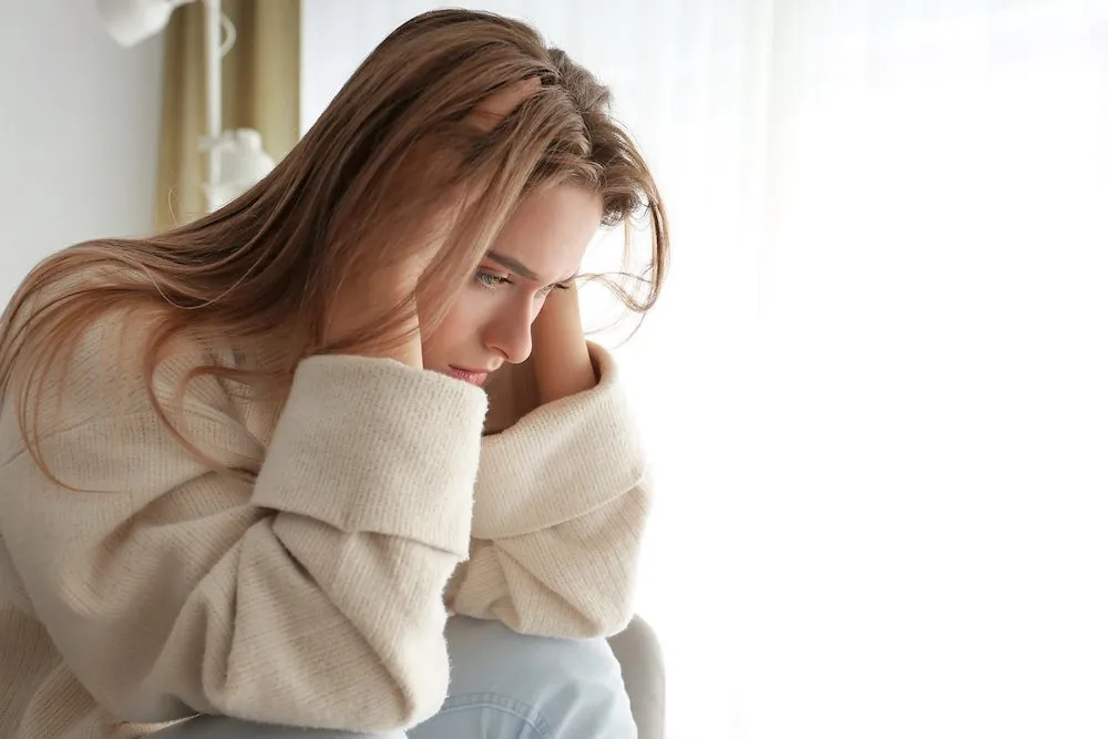 A woman in a cream sweater sitting by a window, looking tired and low