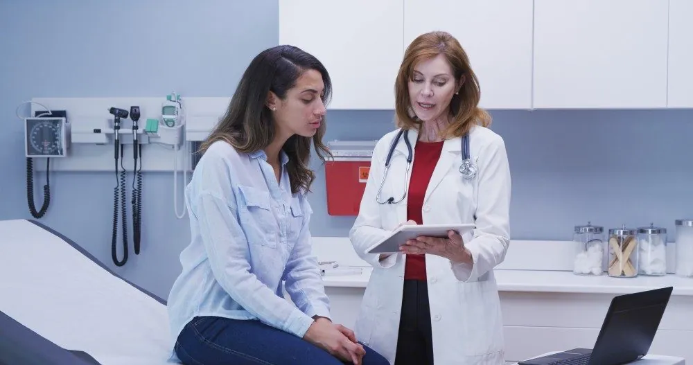 A patient sitting on an exam table while a doctor reviews notes on a tablet