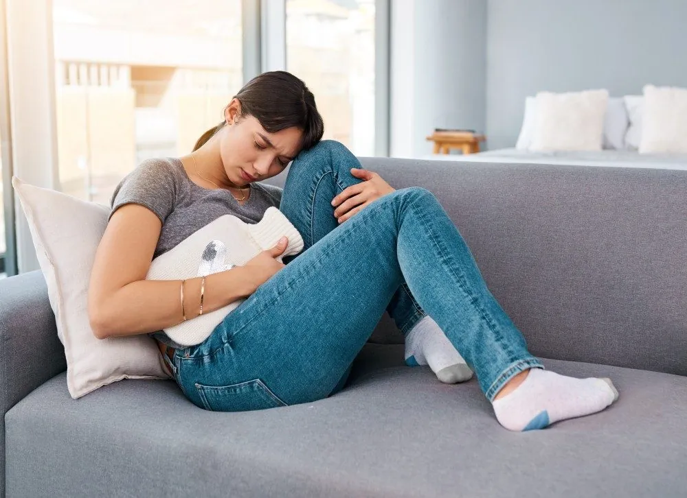 A young woman lying on a sofa hugging a heating pad to her abdomen