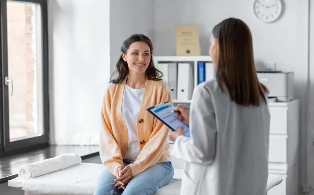 A smiling woman in a consultation room talking with a clinician holding a tablet