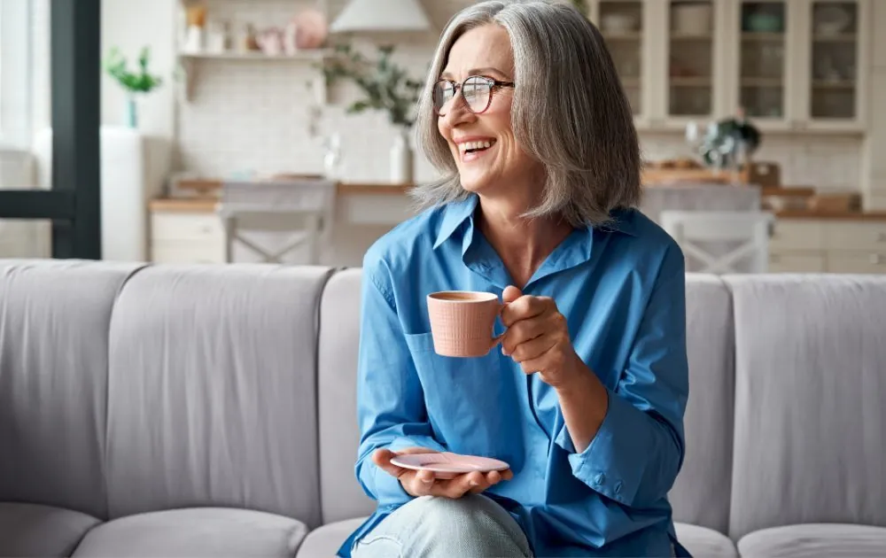 A woman in her fifties smiling with a cup of coffee on her sofa